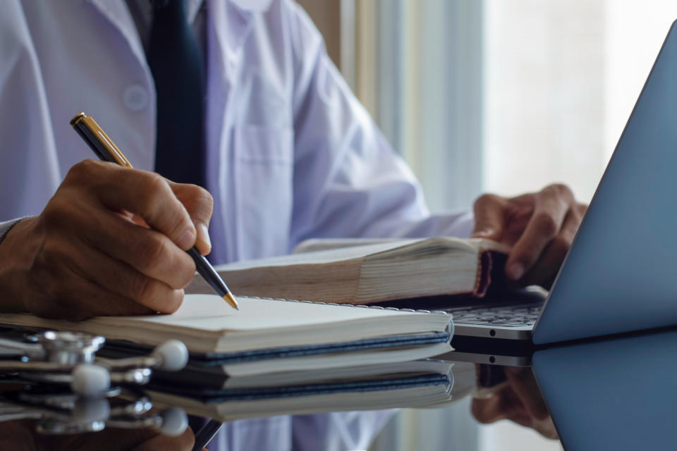 Closeup male doctor or physician reading text book and writing information,work on laptop computer with medical stethoscope on the desk. Online working, telehealth or meditech concept.