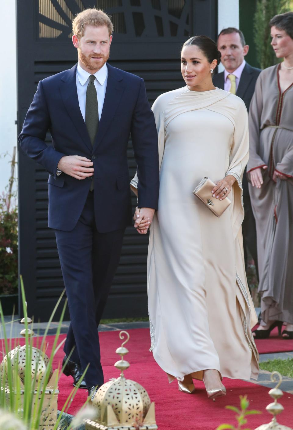 Harry in a navy suit with a white button-down and olive green tie. Meghan is wearing a cream long-sleeve flowing gown with a cape feature that criss crosses on her chest. She's holding a gold clutch and is wearing gold pointed heels.