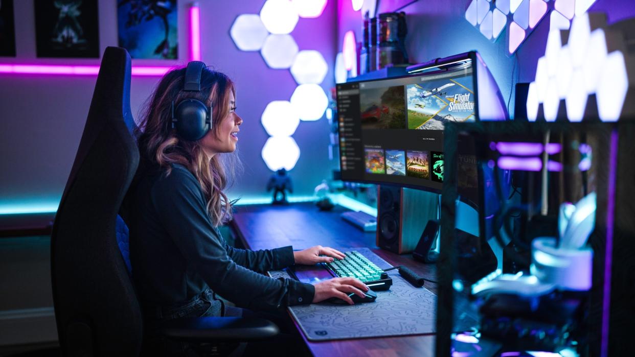  Woman with headphones sitting side on to camera at a desk with keyboard, mouse, and a monitor looking through her PC Game Pass library 