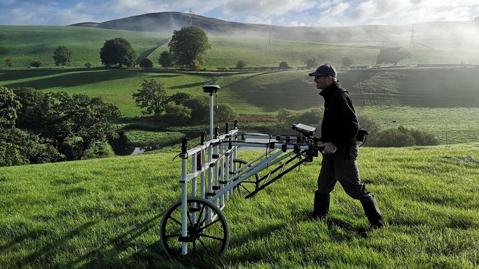 A man dressed in dark colors walks in a grassy field pushing a lawn mower-like instrument for gradiometry.