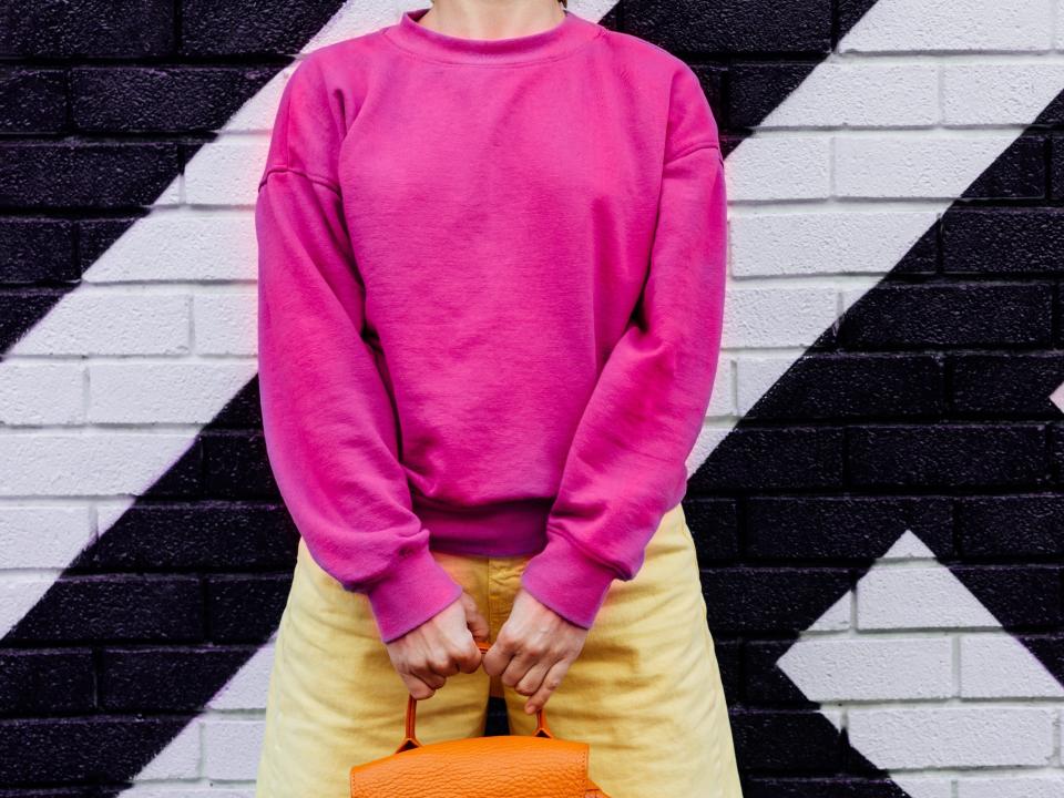 Woman wearing magenta sweater and yellow pants while holding orange bag.