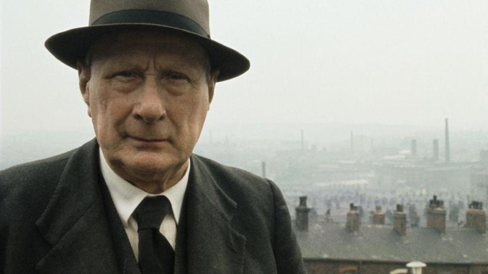 Artist LS Lowry standing in front of his terraced house home in Pendlebury, with the chimneys and factories of Salford in the background