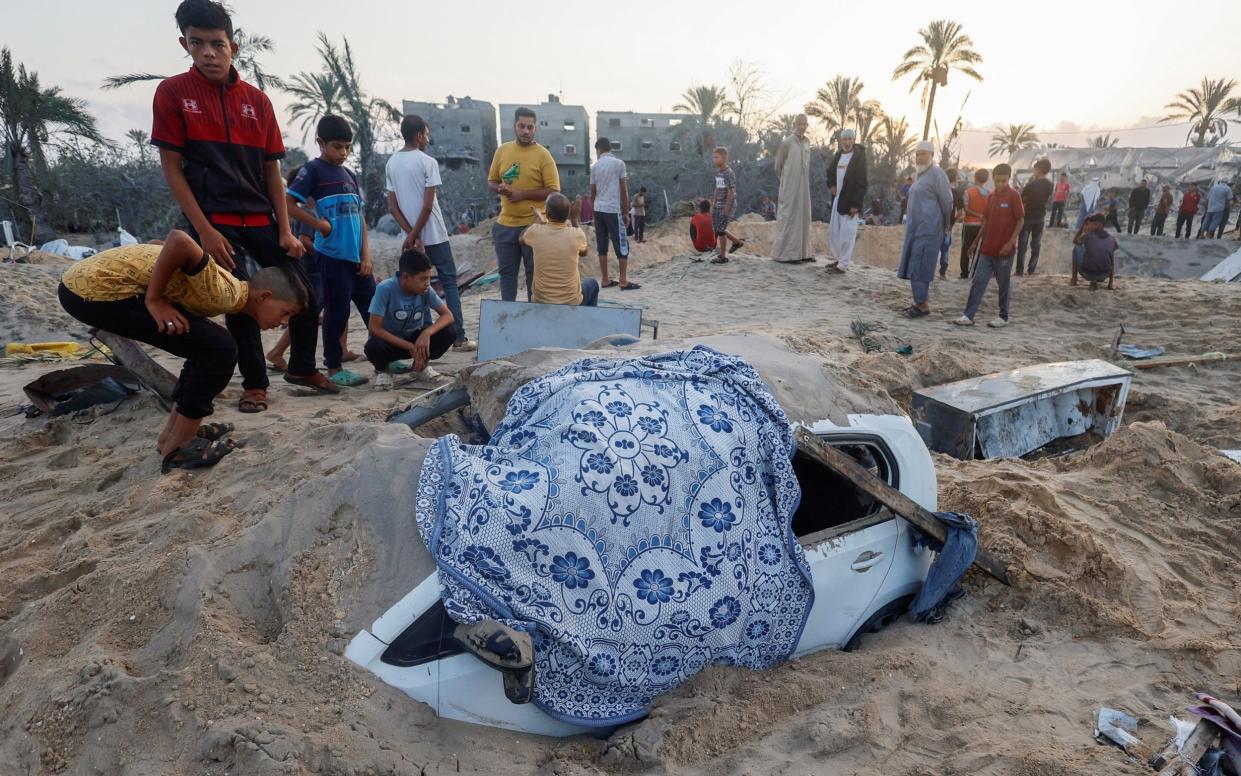 Palestinian boys stand near a buried damaged vehicle at the site following Israeli strikes on a tent camp in southern Gaza