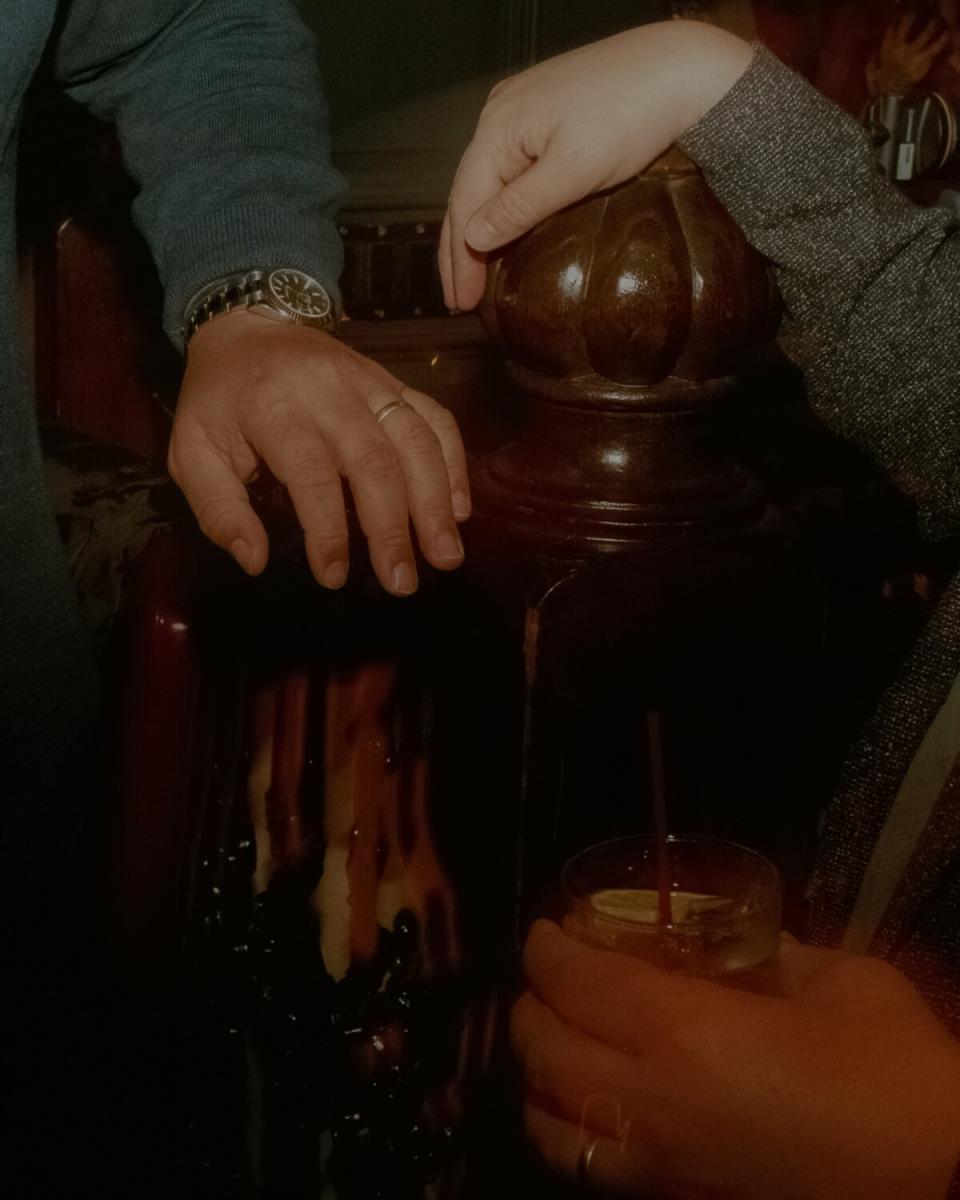 Pairs of hands are seen, one is holding a drinking glass while the other rests on a staircase banister.