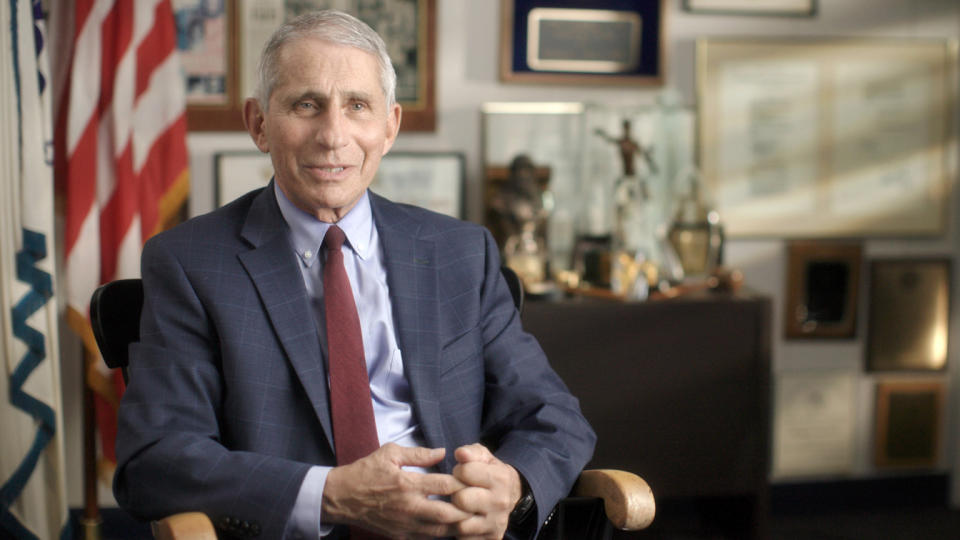 Dr. Anthony Fauci wears a blue suit and dark red tie while sitting in an office in front of plaques, picture frames and an American flag.