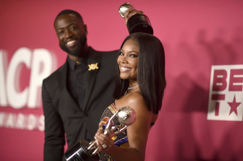 A man in a black suit and a woman in a gold dress smiling and holding gold trophies in front of a red background