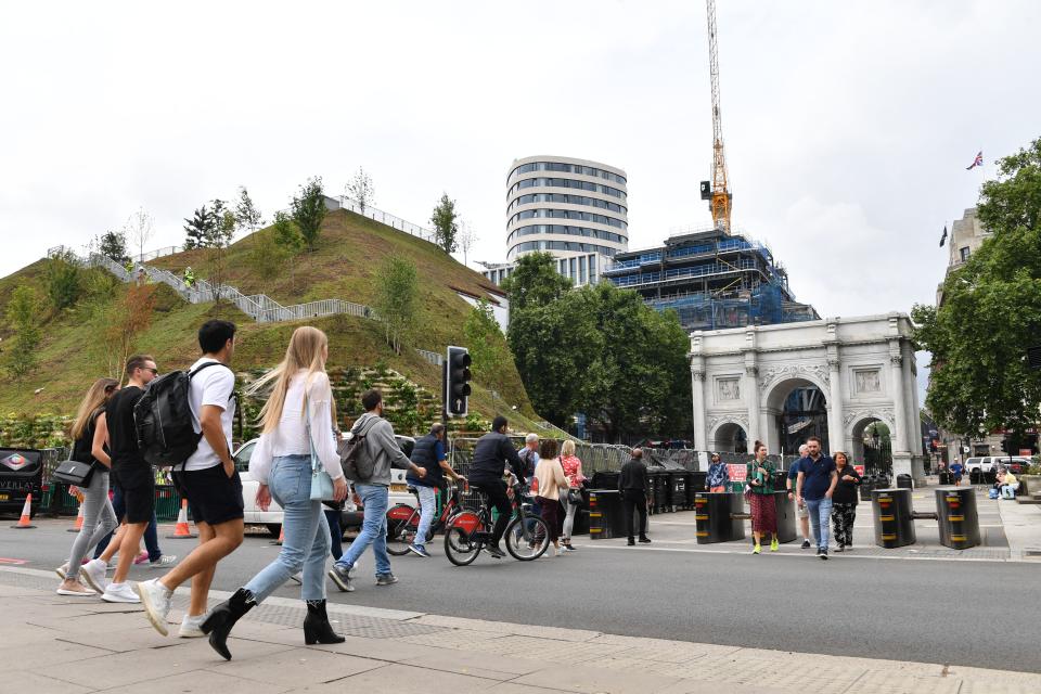 People cross the road next to the Marble Arch Mound, a new temporary attraction, next to Marble Arch in central London on July 25, 2021. - Designed by Dutch architecture company MVRDV, the 25-metre-high landscaped Marble Arch Mound consists of a stairway leading through trees and greenery to a viewing platform at the top. The ticketed new attraction opens to the public on July 26. (Photo by JUSTIN TALLIS / AFP) (Photo by JUSTIN TALLIS/AFP via Getty Images)