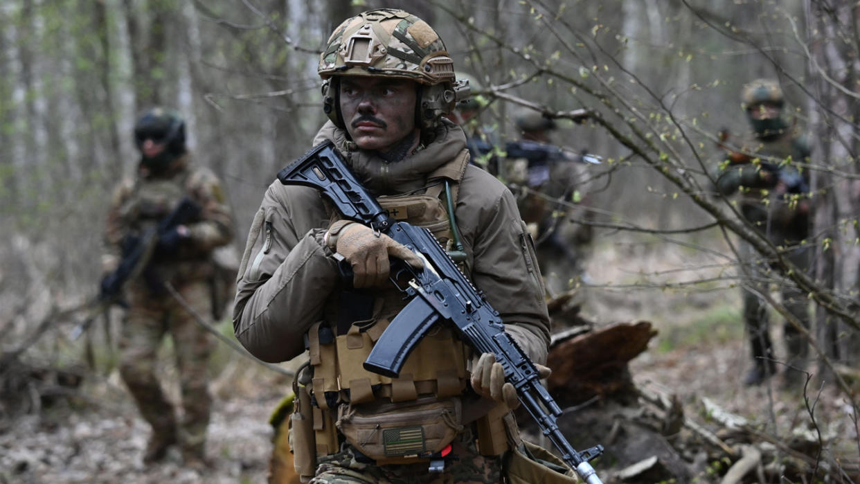 Ukrainian soldiers holding weapons walk through a wooded area.