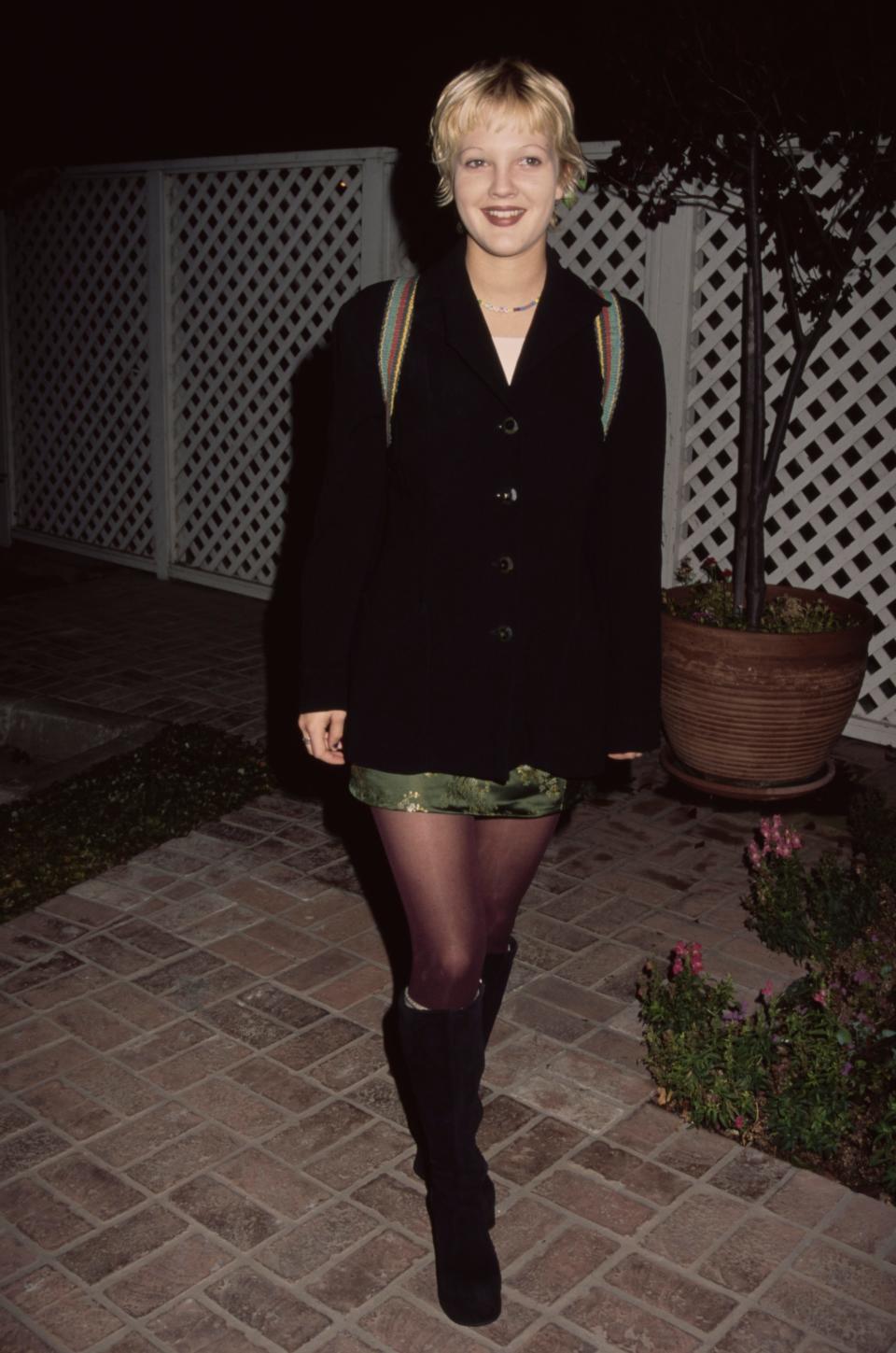 Drew Barrymore, wearing a black jacket over a green miniskirt, with what are possibly shoulder straps of a backpack over the jacket, and black knee-high boots, smiling as she attends an unspecified event, location unspecified, 1994