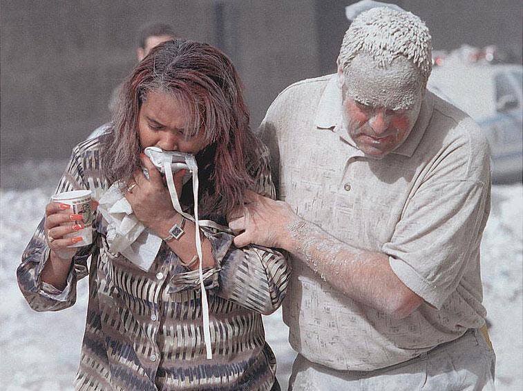 Man covered with ashes assisting a woman walking and holding a particle mask to her face, following the attack (Don Halasy)