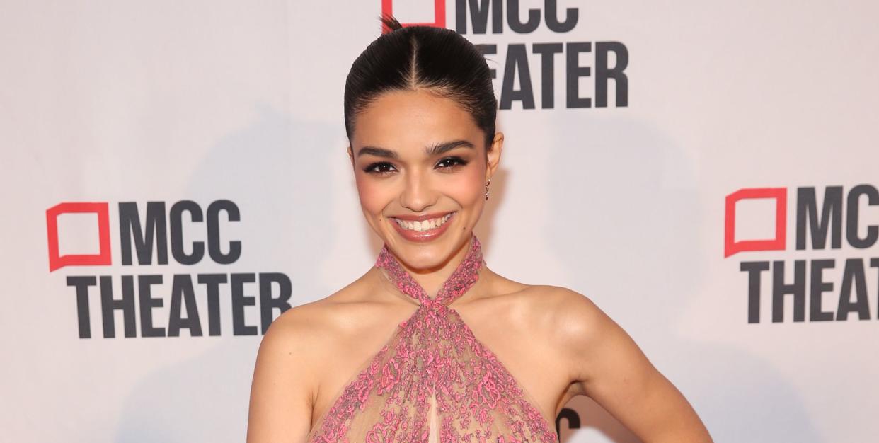 rachel zegler, a young woman stands smiling at the camera with left hand on her hip, she has black hair tied back in a bun and wears a pink lace halterneck dress