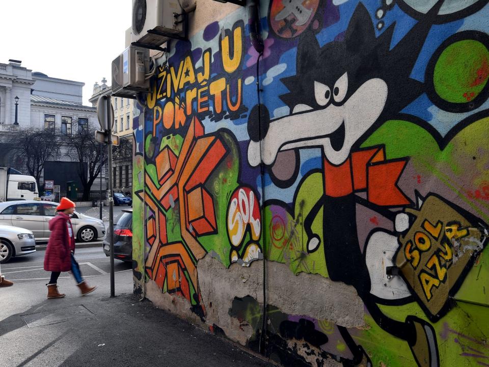 Pedestrians walk past graffiti depicting the official olympic mascot "Vucko" from the XIV Winter Olympic Games held in Sarajevo in 1984, on a painted wall painted mural in a an alley, in Sarajevo city center, on February 7, 2024.