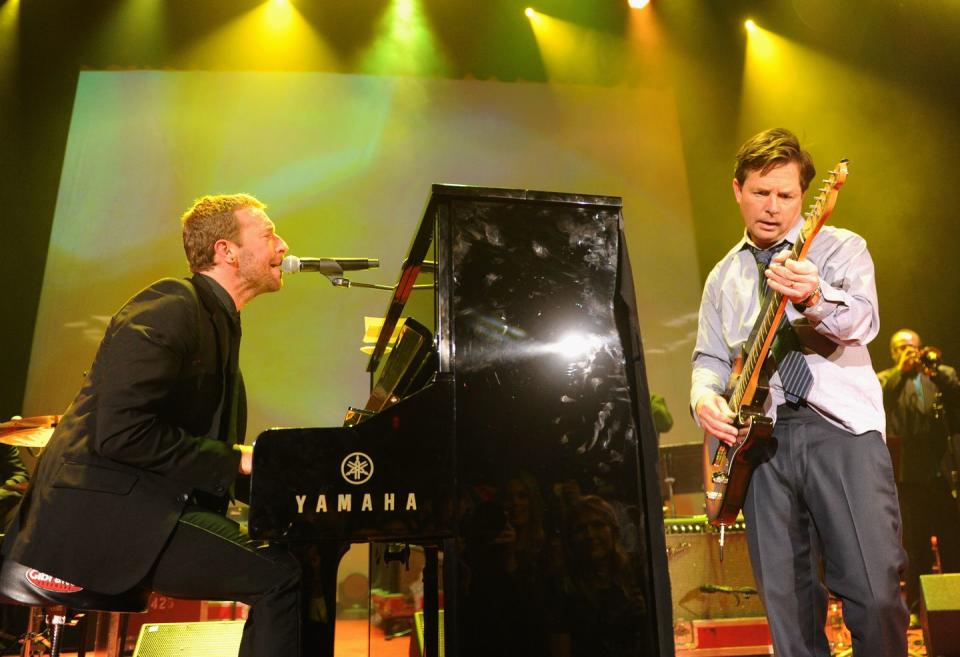 chris martin plays a black upright piano and sings into a microphone while michael j fox stands to the right and plays electric guitar