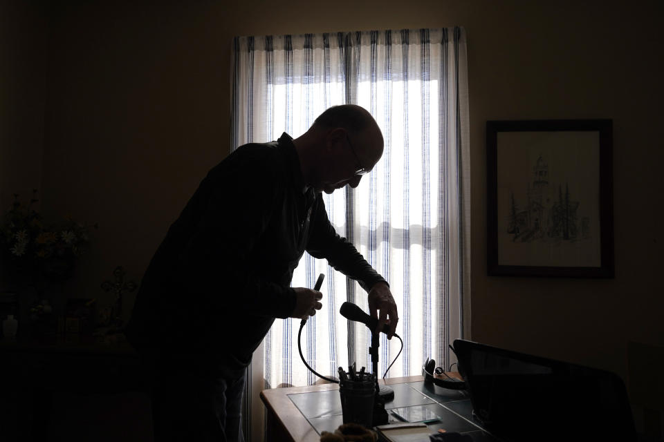 Roger Strukhoff 67, stands in silhouette cleaning off his home office desk Thursday, Jan. 20, 2022, in his DeKalb, Ill., home. Strukhoff was being treated for intestinal bleeding at a hospital outside Chicago this month when he suffered a mild heart attack. Normally, the medical staff would have sent Strukhoff to the intensive care unit, but, overrun with COVID-19 patients, the staff instead had to wheel a heart monitor into his room and quickly administer nitroglycerin and morphine. (AP Photo/Charles Rex Arbogast)