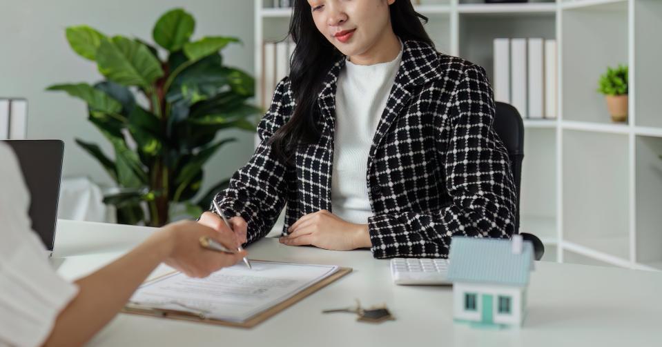 Two women sit at a desk in an office, signing documents. One woman wears a checked blazer, the other is partially visible from behind. Shelves with decor items in the background