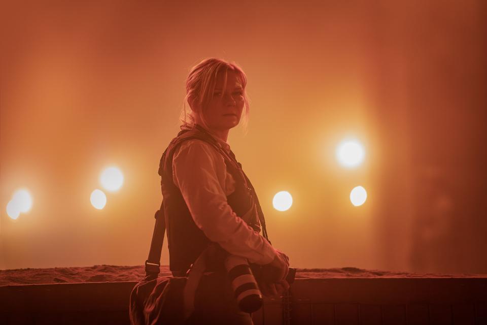 a woman (kirsten dunst) with a camera stands in front of car headlights in a hazy orange scene