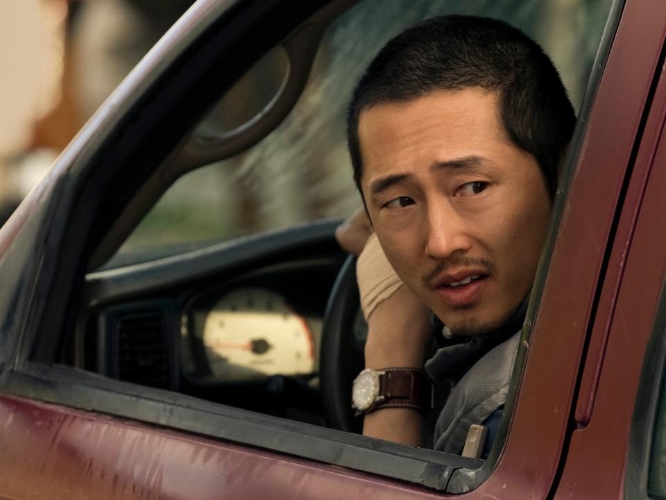 steven yeun as danny cho in beef. he's a man with short-cropped black hair, looking backwards out of the drivers' side window of his red truck with a concerned expression on his face