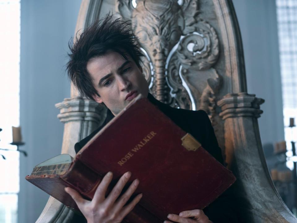 tom sturridge as dream in the sandman, a young man with wild brunette hair, sitting on a stone throne and holding a large, red tome with the text "rose walker" on the front. his hands are splayed across the back of the book and he is reading it intently