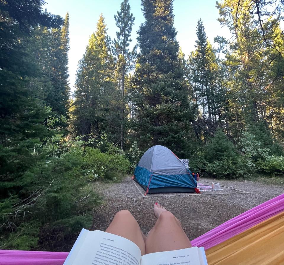 Emily's legs hanging off of a pink and orange hammock, with a book on top of her legs, and a tent and trees in front of her.