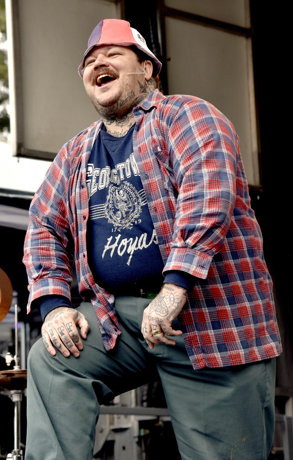 A heavily tattooed Matty Matheson at a cooking demonstration wearing a Georgetown Hoyas t-shirt, a plaid shirt over that, and pants. He's also wearing a bucket hat