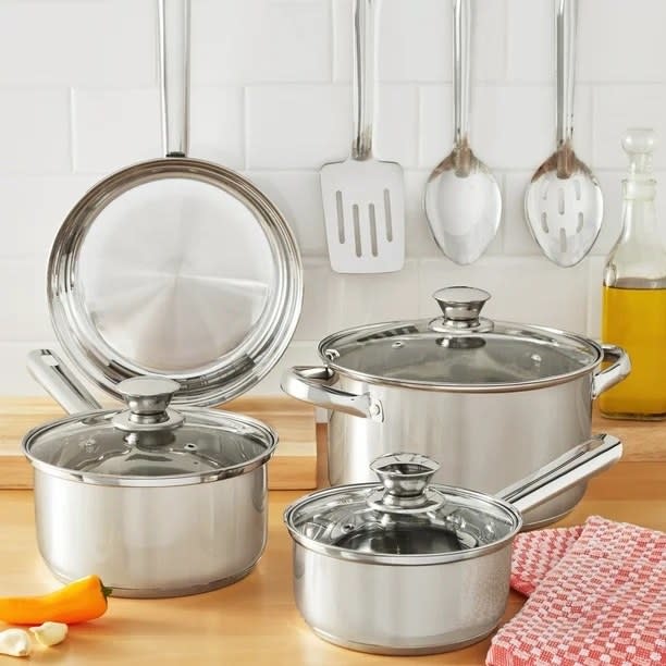 Stainless steel cookware set including pots and pan with lids, displayed on a kitchen counter with utensils hanging in background
