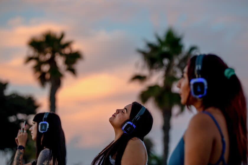 Side view of three people standing with headphones and their eyes closed, against a sunset and palm tree background.