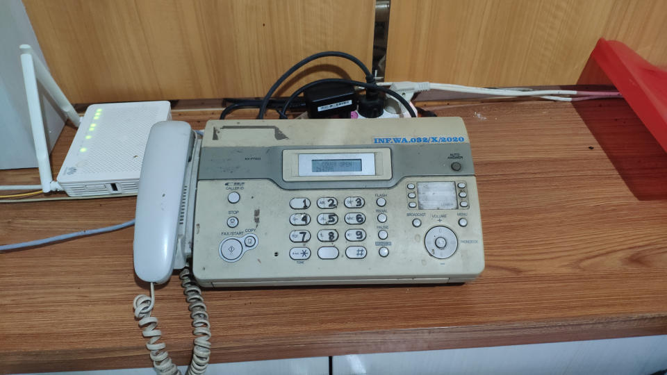 A dusty old fax machine with buttons and a display screen, sitting on a wooden desk next to a modem