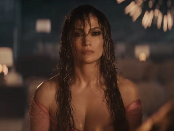 jennifer lopez, sitting in front of a fire, her hair wet and a dress clinging to her skin as fireworks reflect in the background