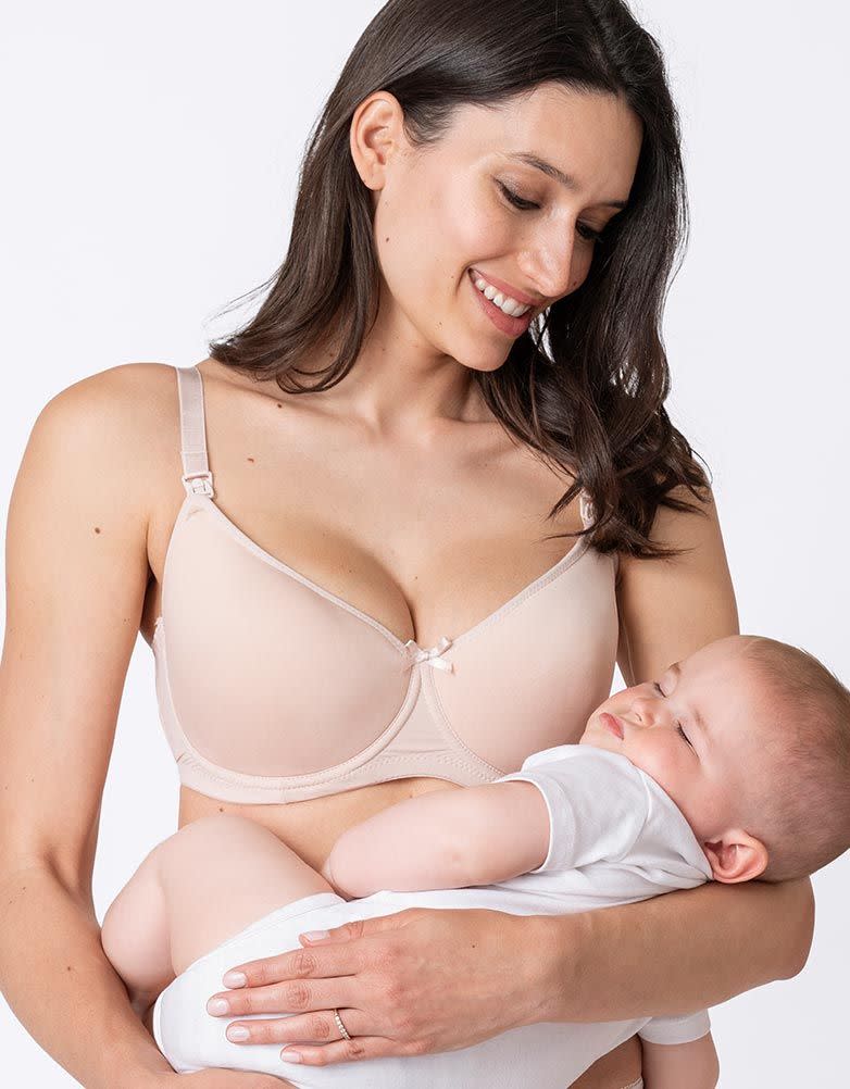 A dark-haired woman in a beige maternity bra smiles down at a sleeping baby in her arms. The baby is in a white suit.