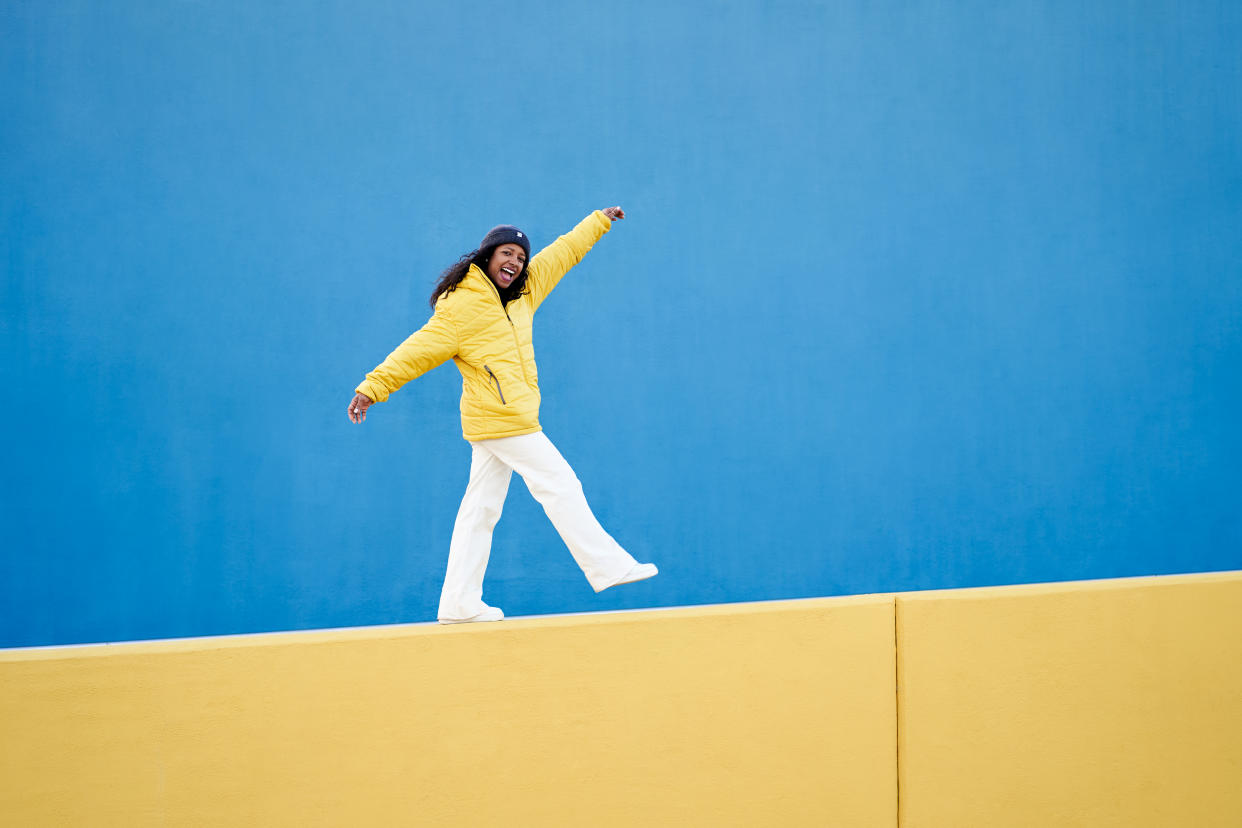 Woman in yellow jacket and white pants balancing on yellow wall with blue background