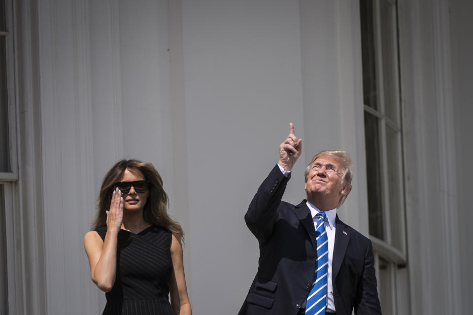 Donald Trump squints and points up at the sky as Melania Trump looks elsewhere with one hand to her sunglasses.