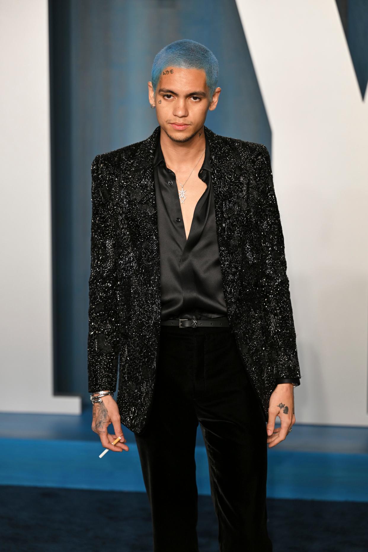 Dominic in a black satin unbottoned top, black sparkly blazer, and black trousers. He has a blue dyed buzz cut an is holding a cigarette on the red carpet.