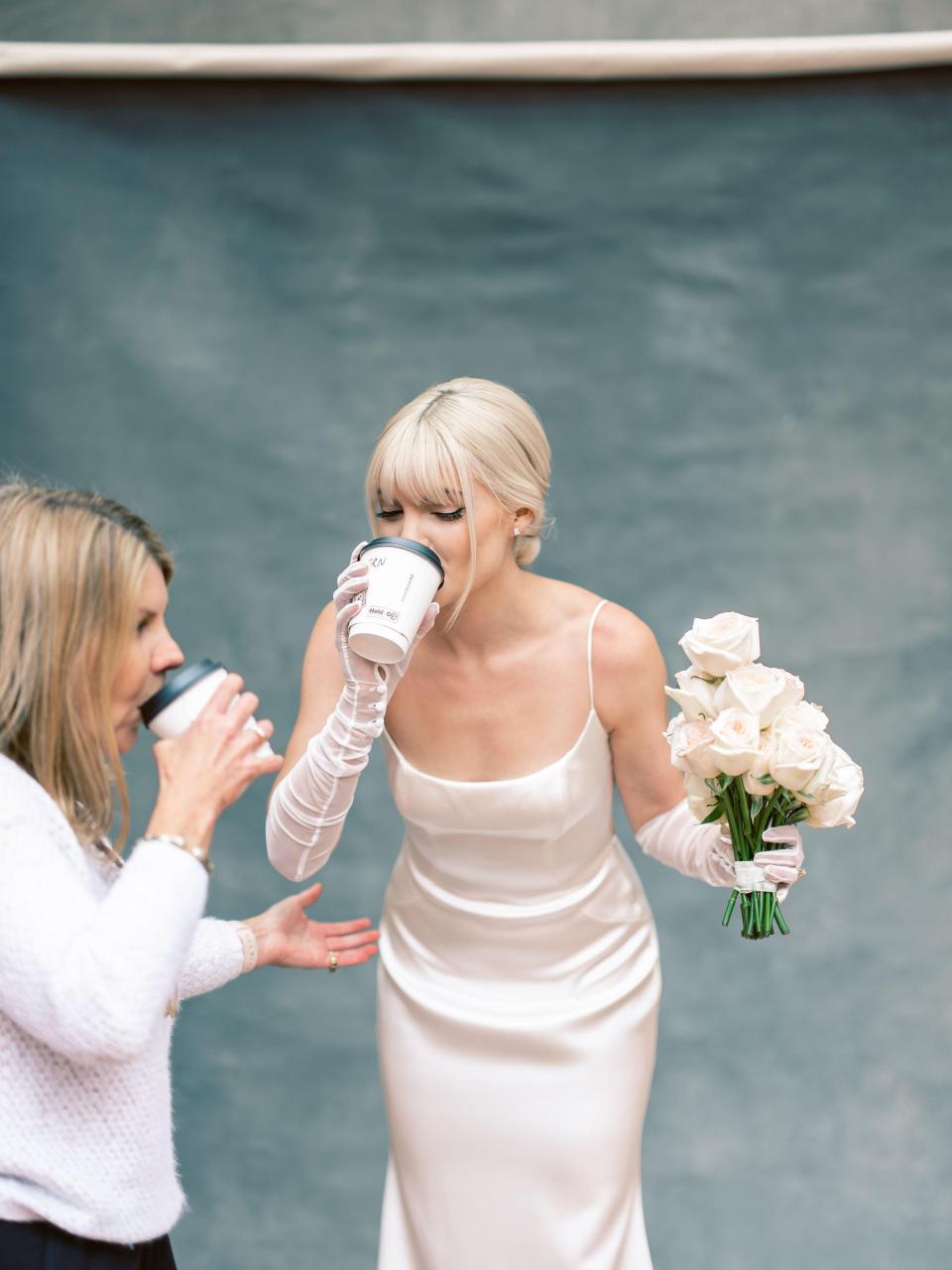 A bride drinks a cup of coffee in her wedding dress while holding a bouquet of flowers in her other hand.