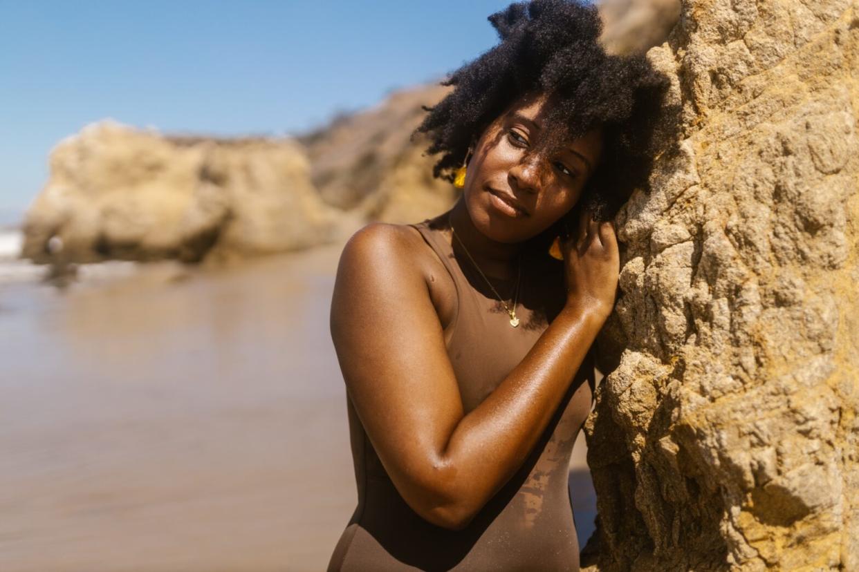 black-woman-in-brown-bathing-suit-leaning-on-a-rock-on-the-beach