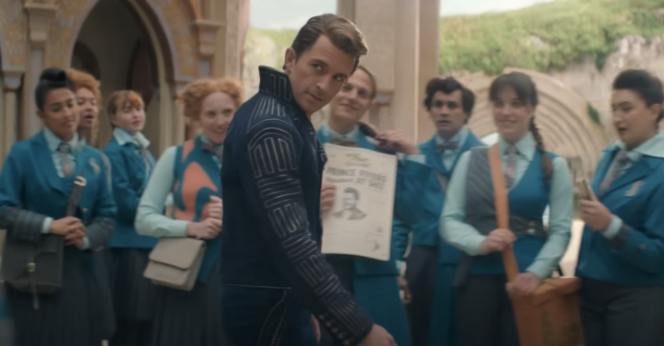 Nicholas Galitzine, wearing a dark suit with intricate patterns, holds up a newspaper, surrounded by eight students in blue uniforms near a stone building