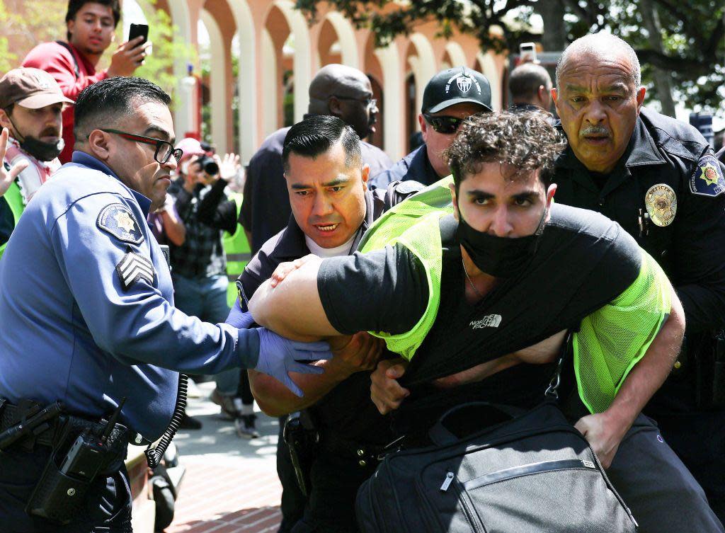 Police officers hold a man wearing a mask and hi viz jacket at a protest on USC