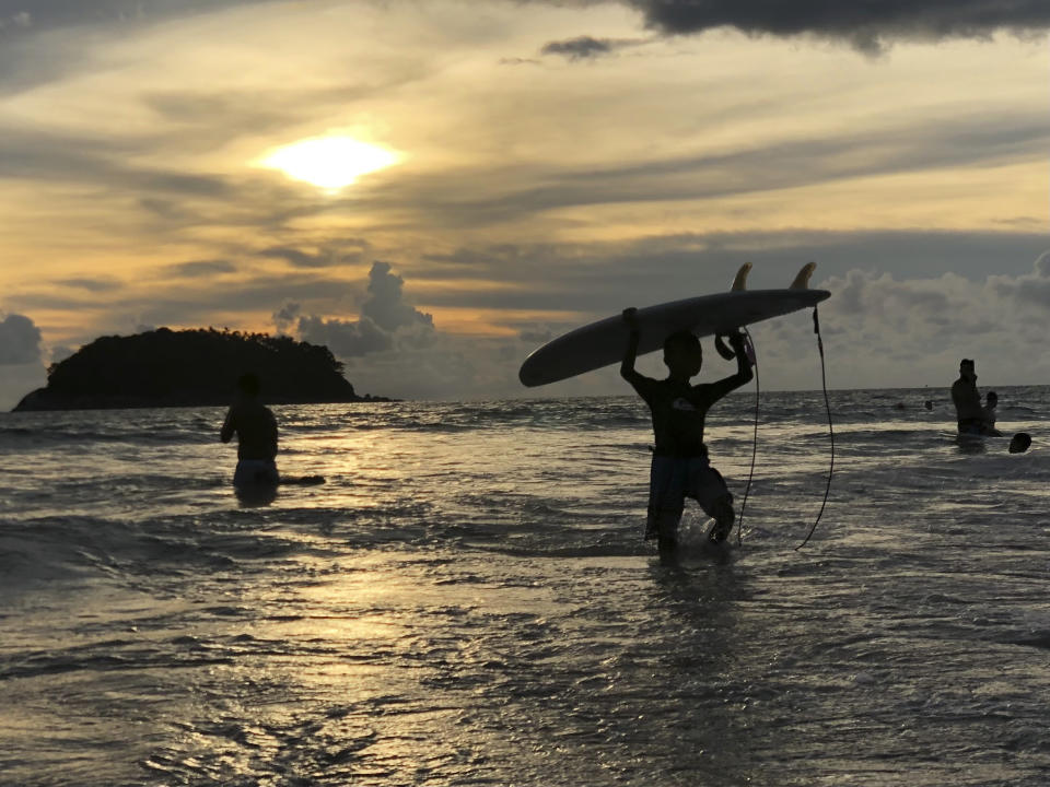 A surfer carries his board ashore as the sun sets over Kata Beach on the resort island of Phuket, Thailand on Sunday, May 26, 2019. Thailand plans to allow vaccinated foreigners to visit the southern resort island of Phuket without quarantining on arrival in a step toward reviving the country's big but battered tourism industry. (AP Photo/Adam Schreck)