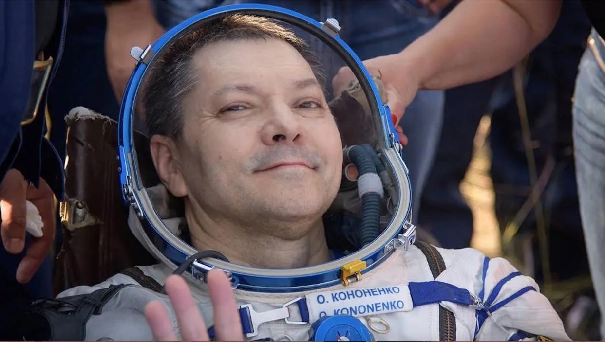  A smiling man in a white spacesuit waves, with several people behind him. 