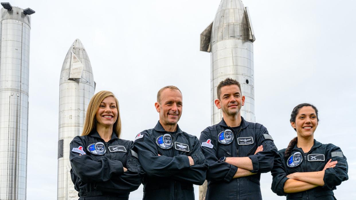  Four people in black flight suits with arms crossed. behind are three silver starship spacecraft. 