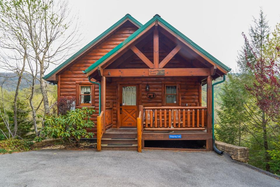 The red wooden cabin with a green roof sits on a road with trees and mountains in the background.