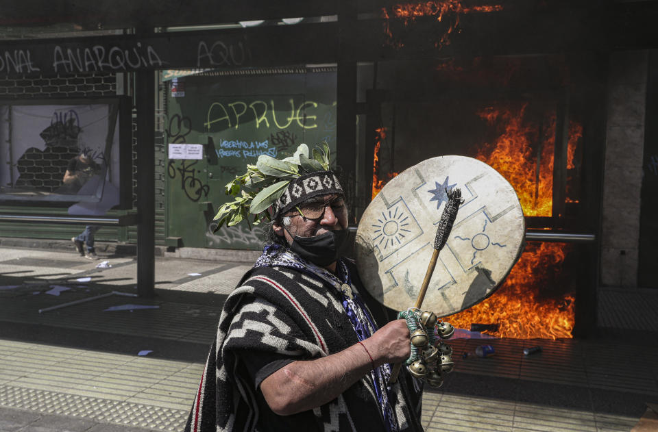 Un indígena mapuche toca el tambor frente a una parada de autobús en llamas durante la tradicional marcha del Encuentro de dos Mundos, que recuerda el descubrimiento de América y la posterior conquista de los pueblos indígenas, en Santiago de Chile el lunes 12 de octubre de 2020. (AP Foto/Esteban Felix)