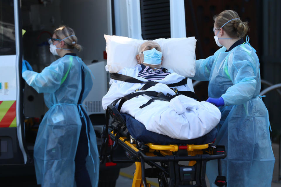 Two nurses wearing protective gear place a male aged care resident into an ambulance. 