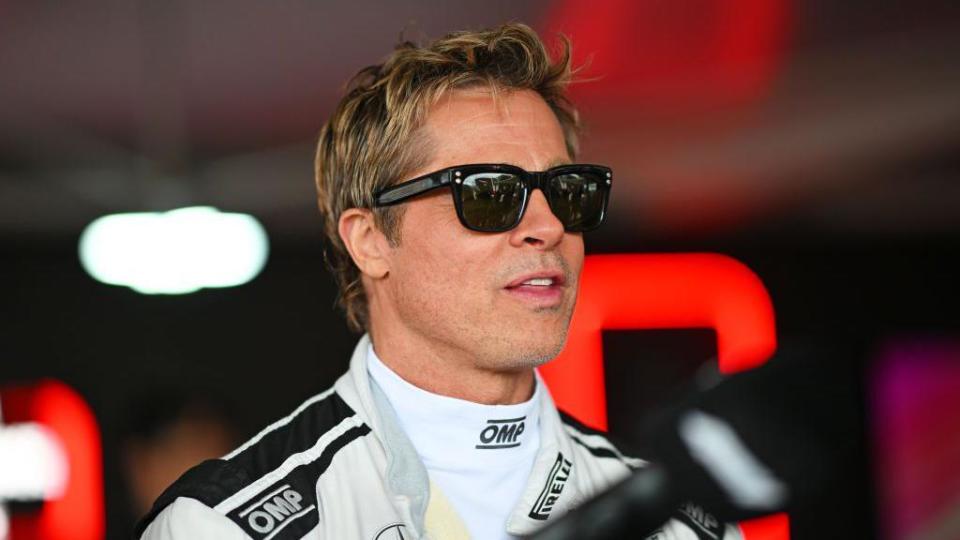 Brad Pitt at a Grand Prix wearing dark glasses and a racing suit