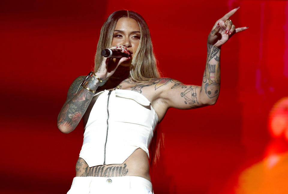 Kehlani performing onstage with a microphone, in a white outfit with prominent tattoos