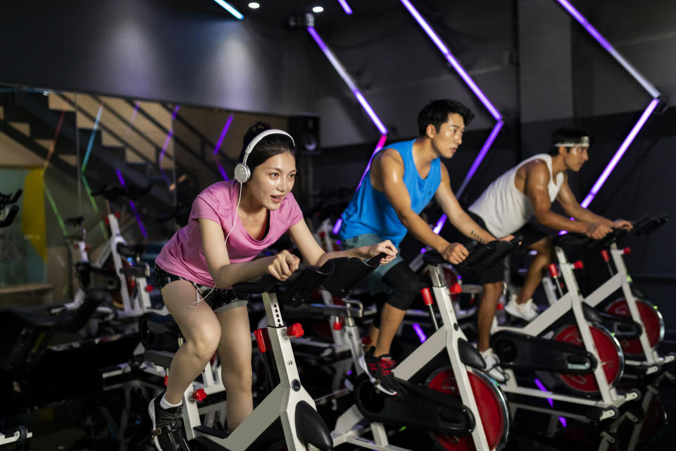 Group of smiling friends at gym exercising on stationary bike. Happy cheerful athletes training on exercise bike. Young men and woman working out at exercising class in the gym.
