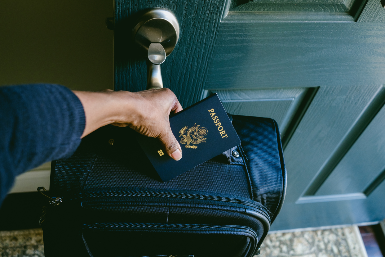 Female hand holding a passport on top of luggage in the front doorway of a home with front door opened to the outside, blurred background of carpet on hardwood floor and a grey wall