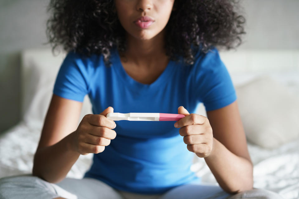 Woman holding a pregnancy test while sitting on a bed. The test has a visible line indicating pregnancy, and she appears contemplative