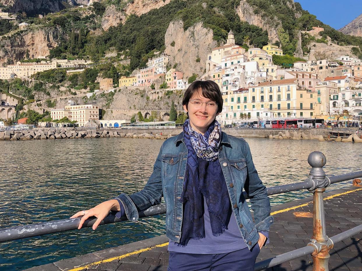 Laura smiles near the water on the Amalfi Coast. There are buildings in the mountains behind her.