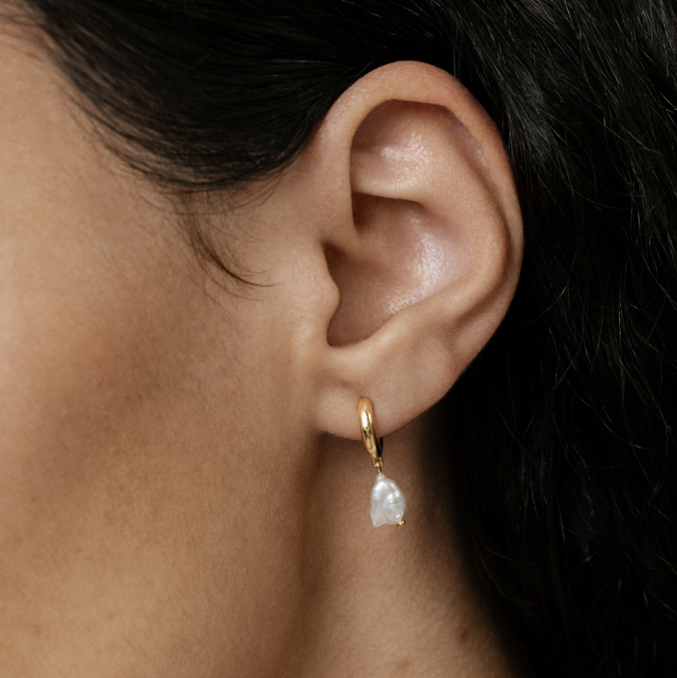 close up of woman with dark hair and gold/pearl Nura Keshi Pearl Huggie Earrings (photo via Monica Vinader)