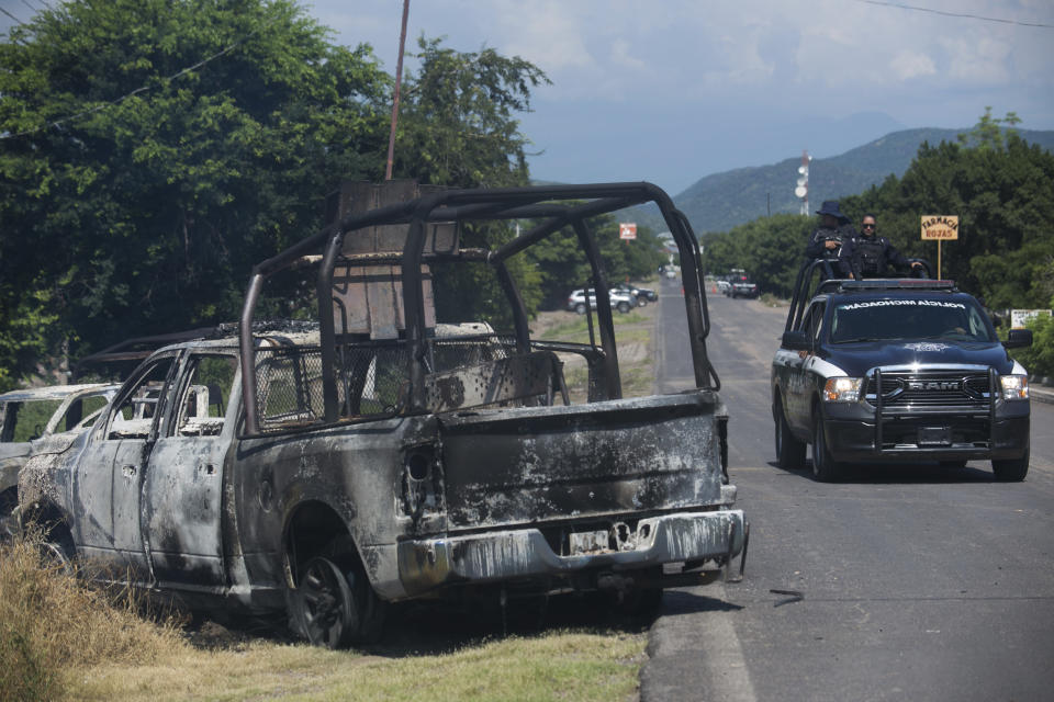 A charred truck that belongs to Michoacan state police sits on the side of the road after it was burned during an attack, as state police drive past in El Aguaje, Mexico, Monday, Oct. 14, 2019. At least 13 police officers were killed and three others injured Monday in the ambush. (AP Photo/Armando Solis)